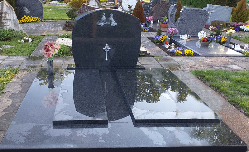 Schwarzer, polierter Grabstein mit Spiegelungen, umgeben von Blumen in einer Friedhofsreihe.