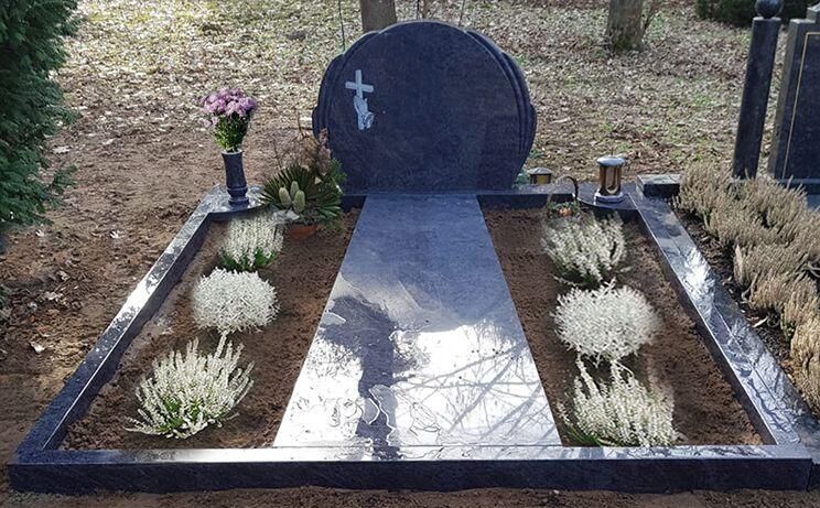 Kleines Gedenkgrab mit schwarzem Grabstein, weißem Kreuz, Blumen und einem polierten Steinweg