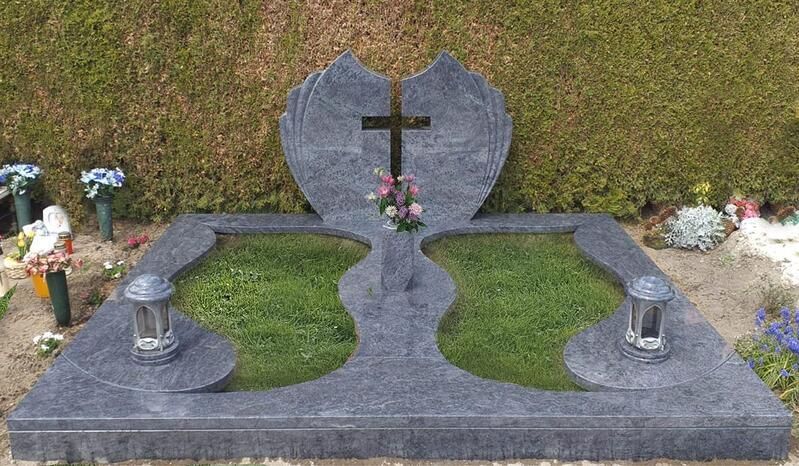 Grabstein mit Kreuz und Blumen, eingerahmt von zwei kleinen Laternen auf einem grasbewachsenen Friedhofsplatz.