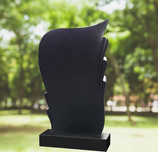 Abstrakte schwarze Skulptur auf einem Sockel im Freien, im Hintergrund verschwommene grüne Bäume.