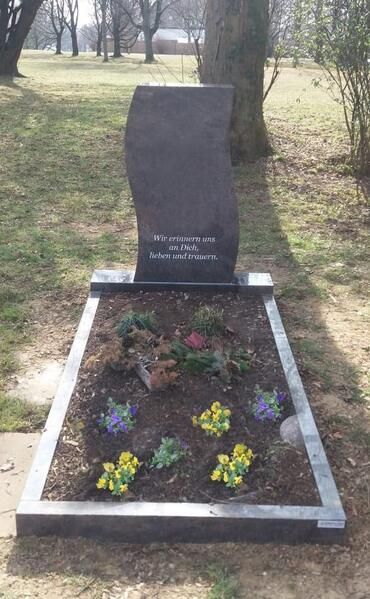 Grabstein mit einem Blumenbeet in einem grasbewachsenen Park, weiß umrandet und mit kleinen bunten Blumen bepflanzt.