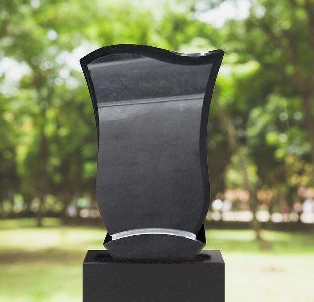 Schwarze Gedenktafel auf einem Sockel im Freien, im Hintergrund verschwommene grüne Bäume.