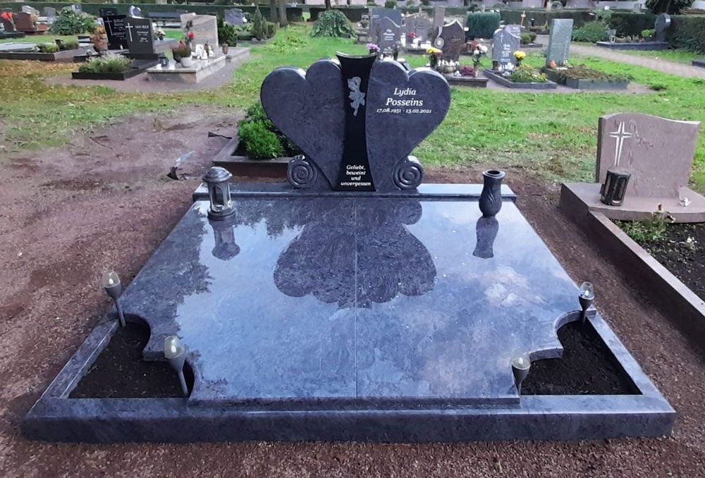 Schwarzer Granitgrabstein mit herzförmigem Grabstein auf einem Friedhof, in dessen poliertem Stein sich Bäume und Himmel spiegeln.