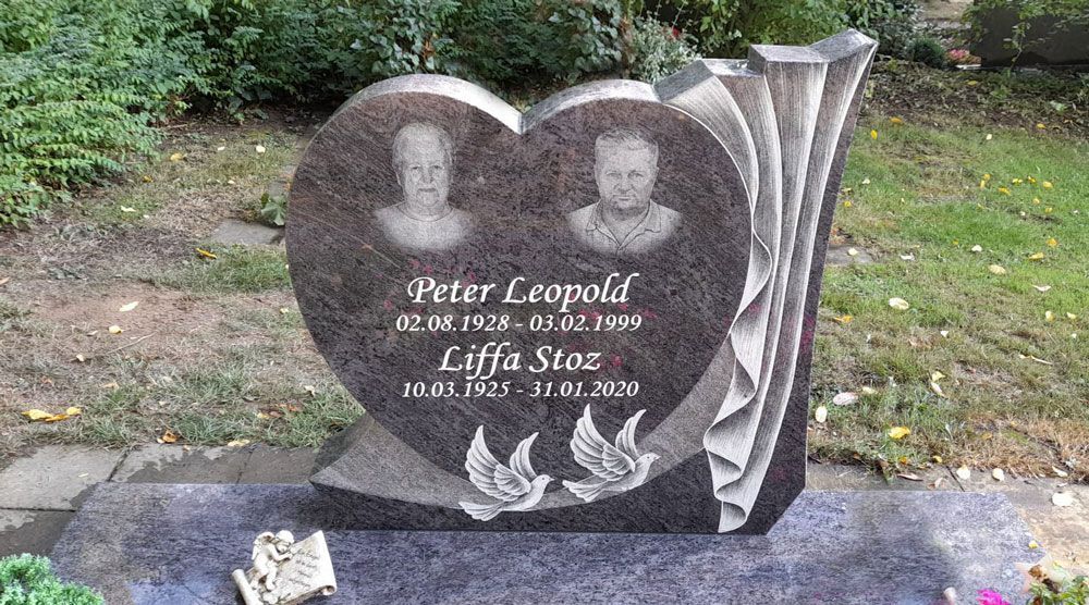 Herzförmiger Granitgrabstein mit zwei Porträtfotos auf einem grasbewachsenen Friedhof.