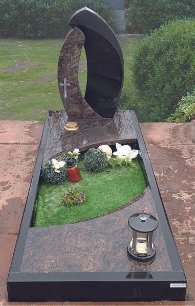 Modernes Grab mit offenem, geschwungenem Grabstein, Blumen, Gras und einer Laterne auf einem Friedhof