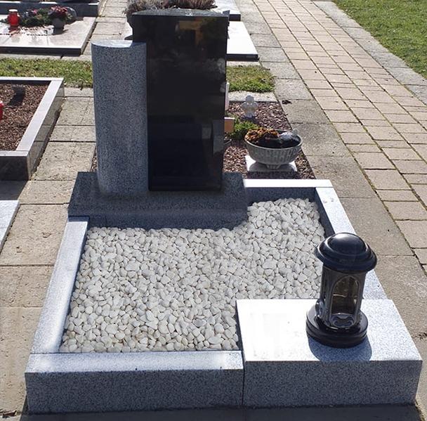 Grabstein mit weißem Kiesrand und Laterne auf einem Friedhofsweg.