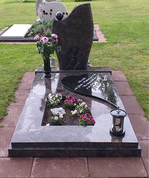 Dunkler Granitgrabstein mit rosa und weißen Blumen auf einem Friedhofsplatz