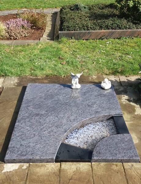 Grabstein mit zwei kleinen weißen Figuren darauf in einem Friedhofsgarten