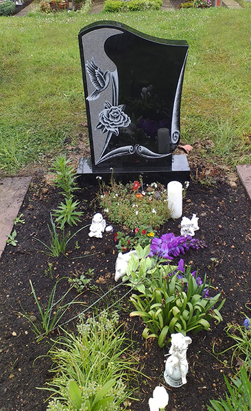 Schwarzer Grabstein mit floraler Gravur, umgeben von Blumen und kleinen Gedenkfiguren auf einem Friedhof