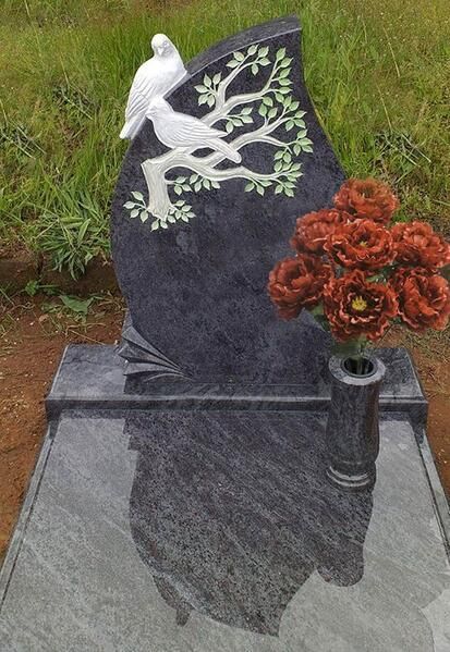 Schwarzer Granitgrabstein mit weißer Taube und floralem Dekor, rechts davon rote Blumen und eine Vase.