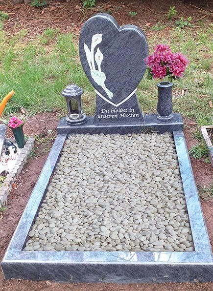 Herzförmiger Grabstein mit weißem Blumendekor, aufgestellt in einem mit Kies gefüllten Friedhofsfeld mit Blumenvasen.