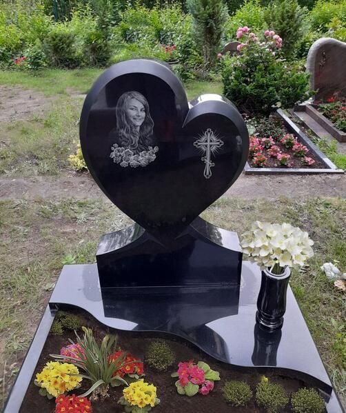 Schwarzer, herzförmiger Grabstein mit Porträt, Kreuz und Blumen in einem Friedhofsgarten