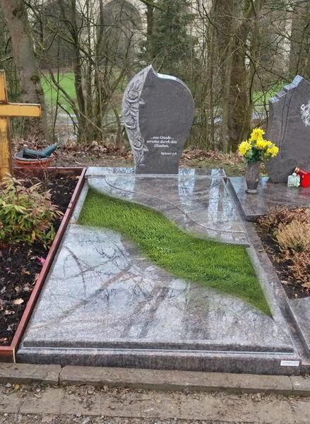 Grabstein in einer landschaftlich gestalteten Friedhofsanlage mit grauem Stein, Rasenstreifen und gelben Blumen