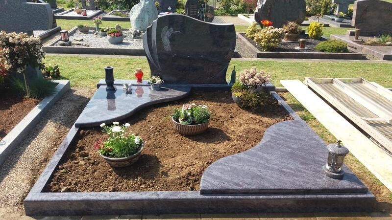 Grabdenkmal mit schwarzem Grabstein, grauem Pflaster, Topfpflanzen und kleinen Figuren auf einem Friedhof