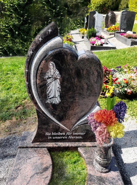 Herzförmiger Grabstein aus schwarzem Granit mit Blumen in einem Friedhofsgarten.