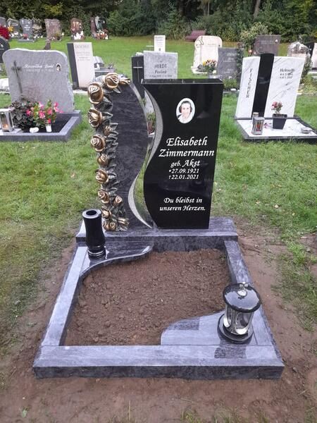Schwarzer Grabstein mit weißer Inschrift und verzierter Seite auf einem Friedhof, in dessen Nähe frische Erde und Laternen stehen.