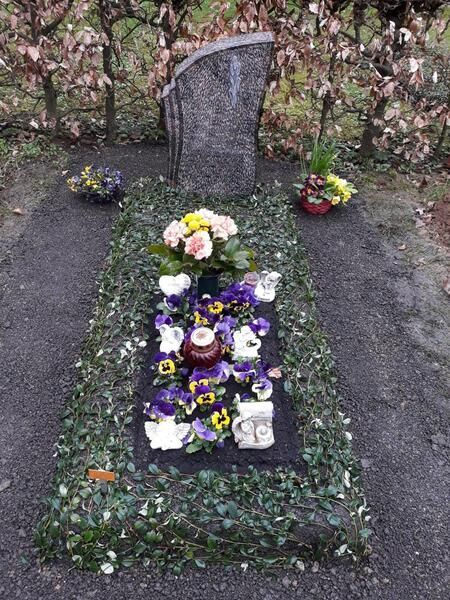 Grabstein, umgeben von Blumenarrangements auf einem Friedhofsgelände.