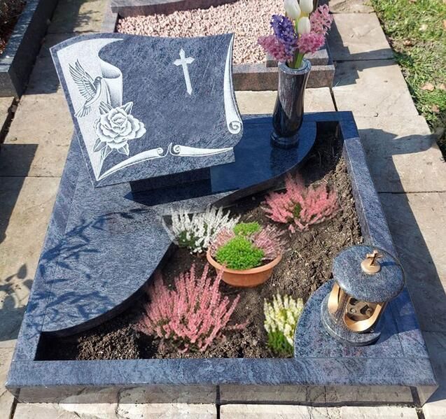 Grabstein mit weißer Rose und Kreuz, umgeben von Blumen und Laternen auf einem Friedhofsplatz