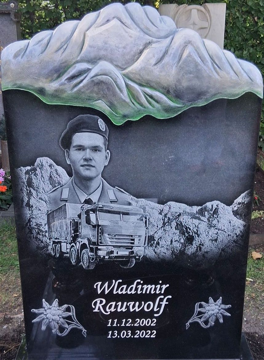 Herzförmiger Grabstein mit zwei Porträtfotos, eingravierten Namen und weißen Blumenverzierungen auf einem grasbewachsenen Friedhof.