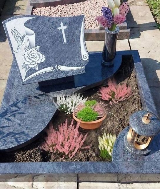 Schwarzer Granitgrabstein mit weißer Blumengravur und Kreuz, umgeben von Blumen auf einem Friedhofsplatz