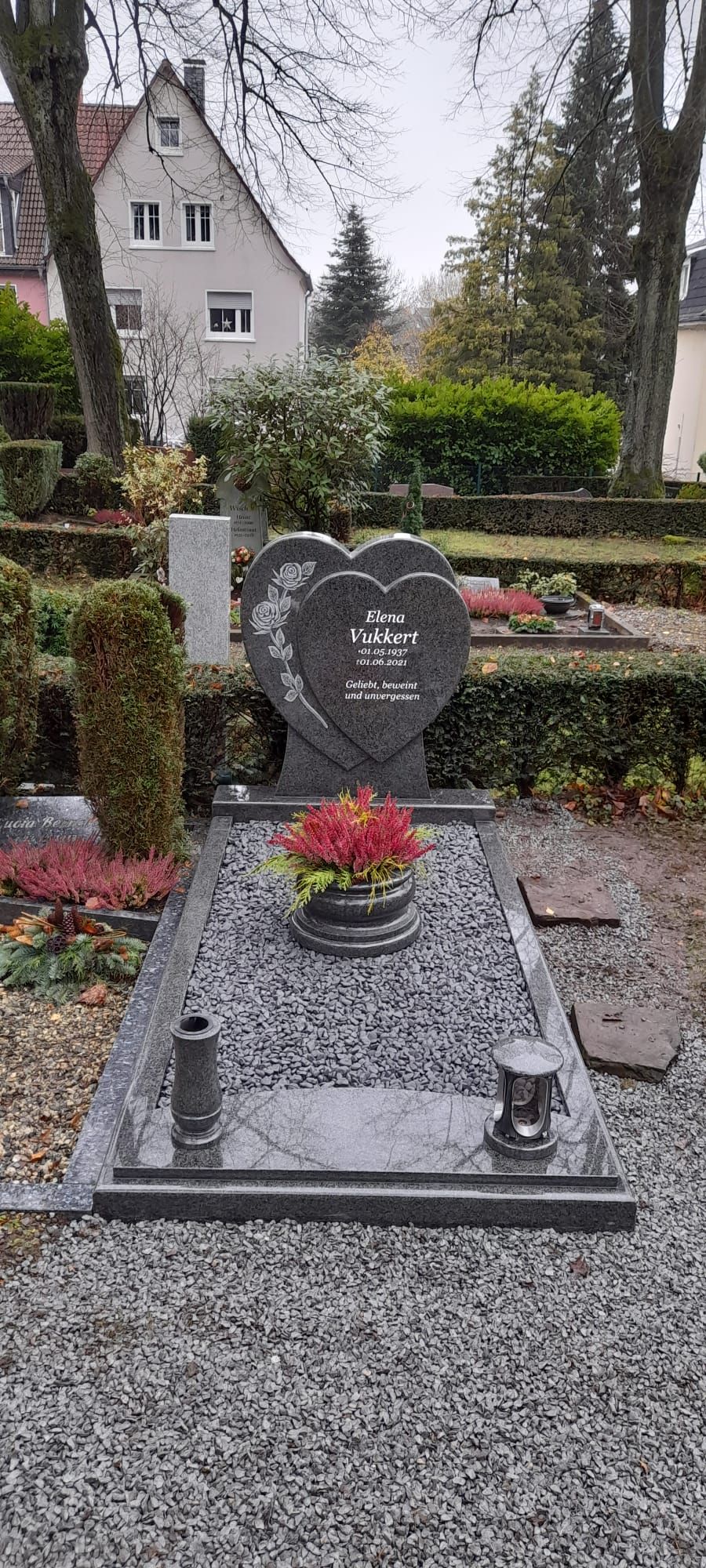 Herzförmiger Grabstein mit roten Blumen auf einem Kiesfriedhof, im Hintergrund ein Haus und Bäume.