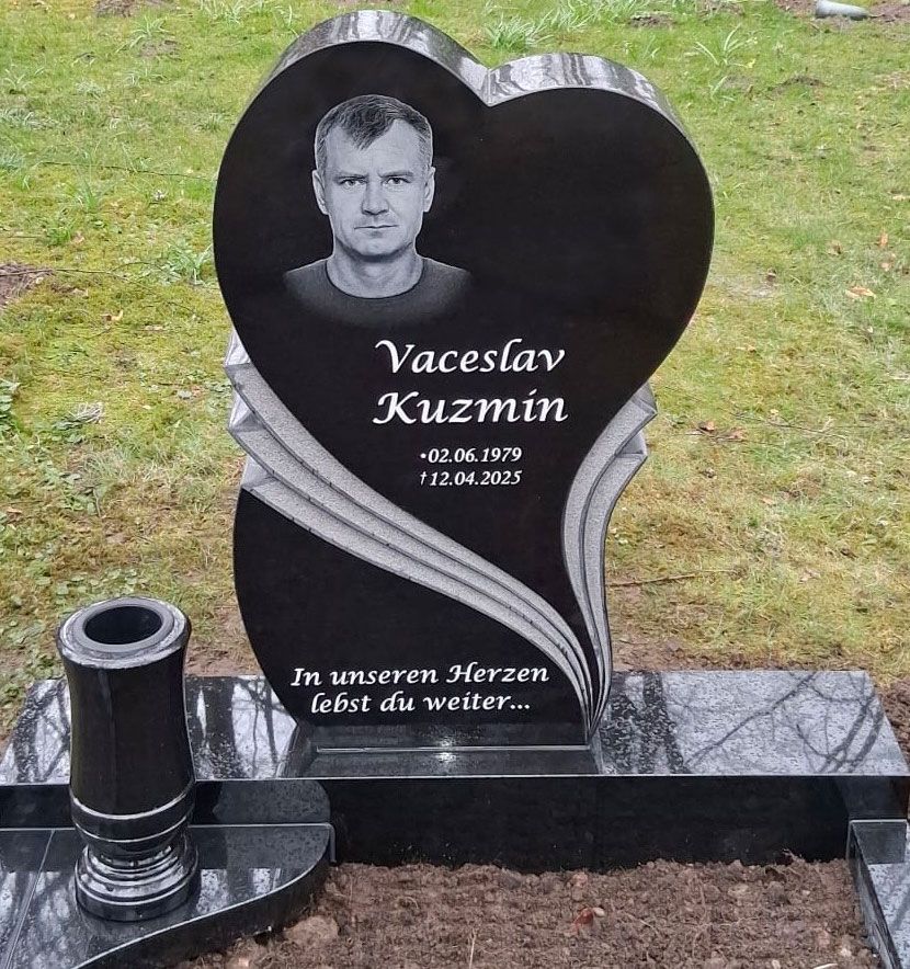 Herzförmiger schwarzer Grabstein mit Porträt, Blumen und deutscher Inschrift auf einer Grabstelle mit Rasen