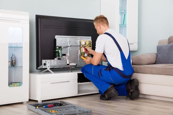 Ein Mann im blauen Overall repariert kniend in einem Wohnzimmer einen Fernseher mit einem Multimeter.