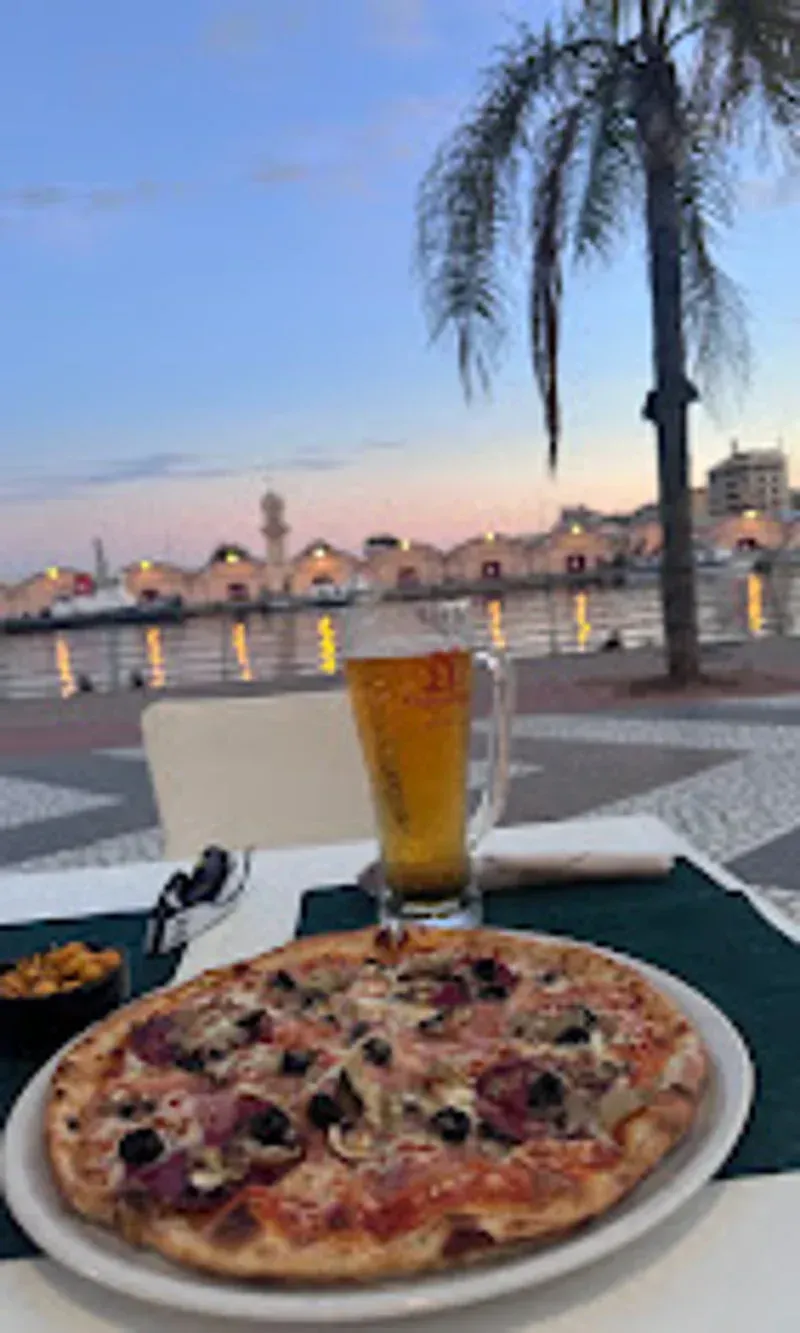 Pizza, cerveza y snacks en una mesa frente al mar al anochecer, con edificios y palmeras al fondo.