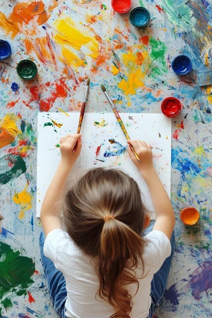 Niña con coleta pintando sobre lienzo, rodeada de manchas de pintura de colores y botes de pintura.