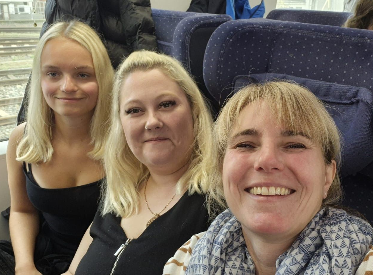 Drei lächelnde blonde Frauen in einem Zug. Eine trägt ein schwarzes Oberteil, die andere einen Schal. Zugfenster sichtbar.