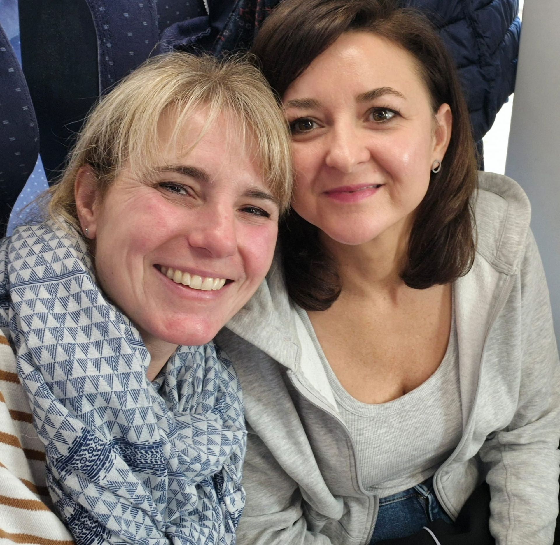 Zwei lächelnde Frauen, eine mit blonden Haaren und einem Schal, die andere mit braunen Haaren.