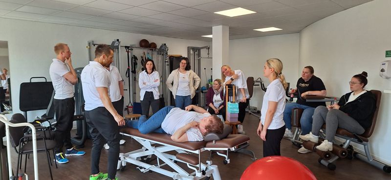 Eine Gruppe von Menschen steht um eine Frau herum, die in einem Fitnessstudio auf einem Tisch liegt.