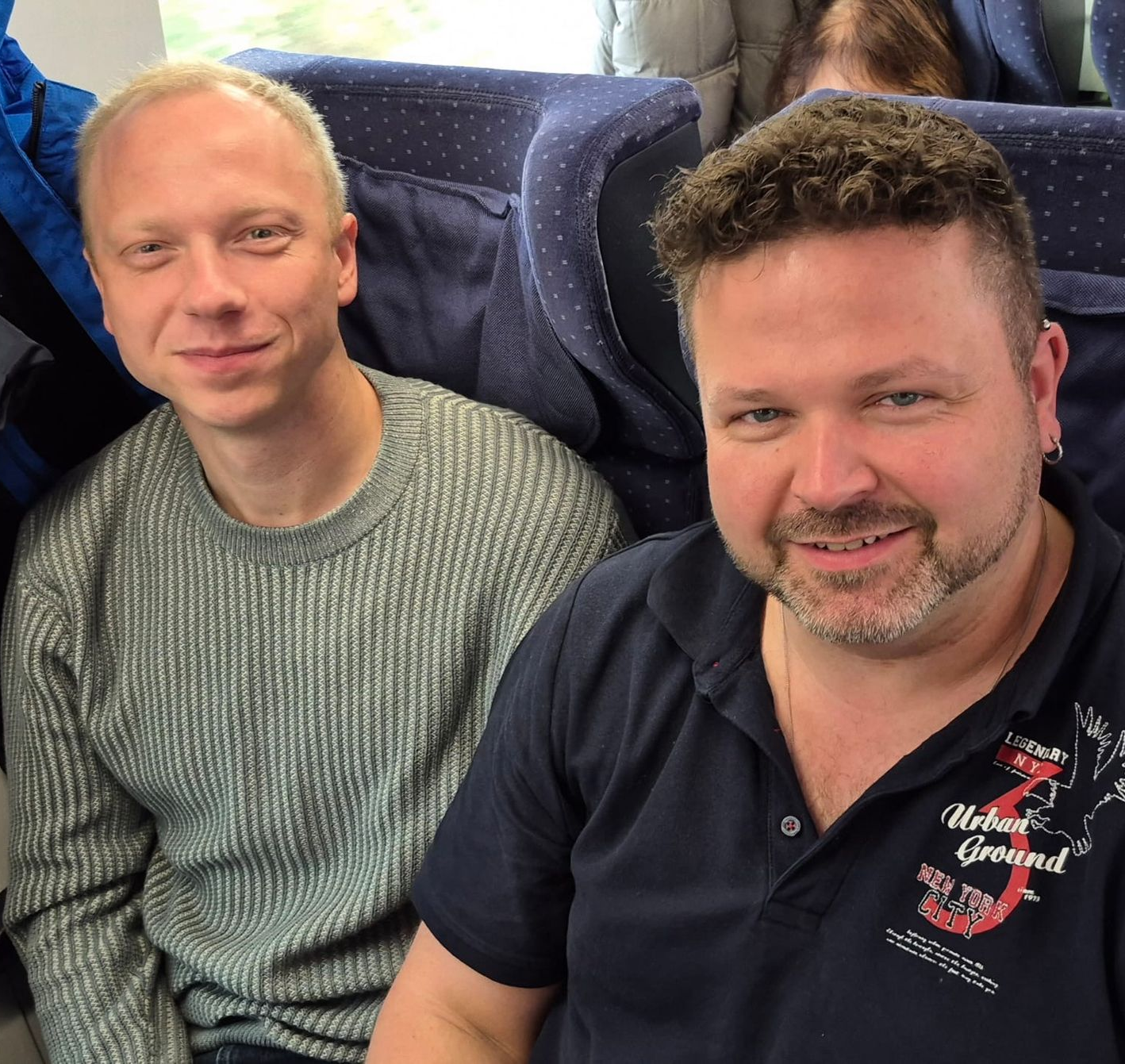 Zwei lächelnde Männer in einem Zug. Einer mit hellem Haar, der andere mit dunklem.