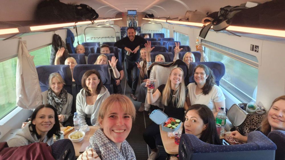 Lächelnde und winkende Menschen in einem Zug, einige halten Essen und Getränke in der Hand.