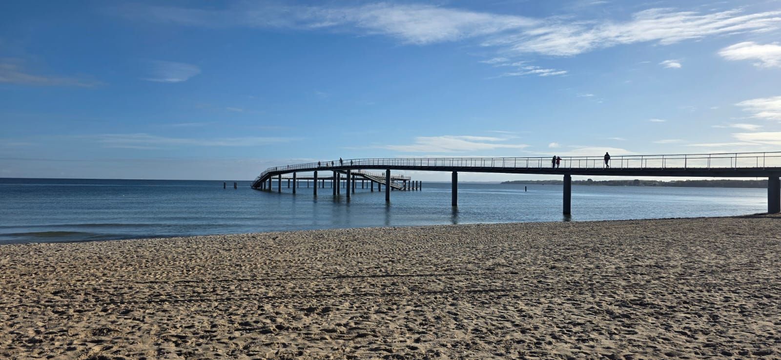 Ein Pier erstreckt sich in den Ozean, auf dem Menschen spazieren gehen, ein sonniger Tag mit blauem Himmel und Strand.