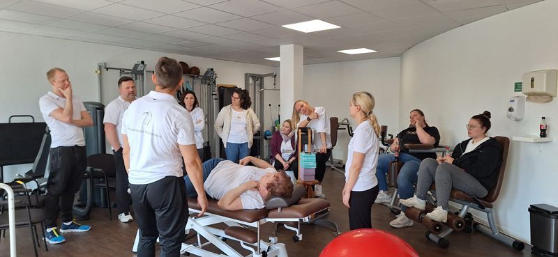 Eine Gruppe von Menschen steht um einen Mann herum, der in einer Turnhalle auf einem Tisch liegt.