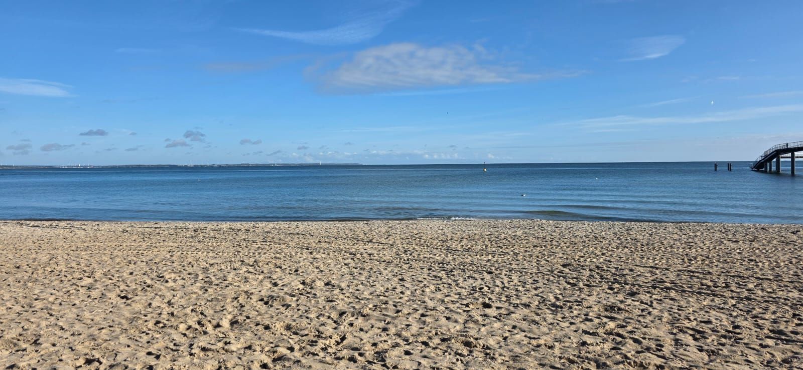 Strand mit blauem Himmel, Sand und Meer. Ein Pier ragt ins Wasser.