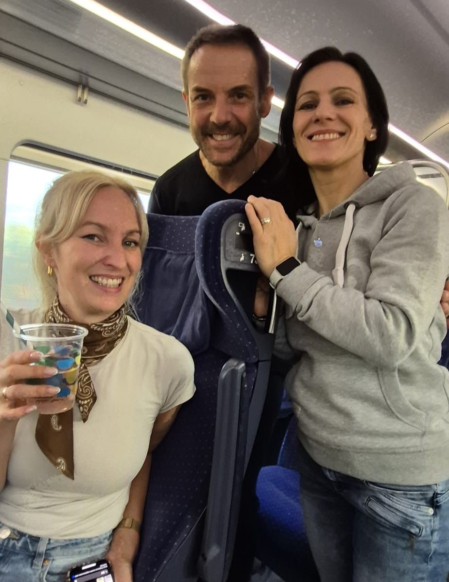 Drei lächelnde Menschen in einem Zug: Frau mit Getränk, Mann mit Schnurrbart, Frau mit grauem Kapuzenpullover.