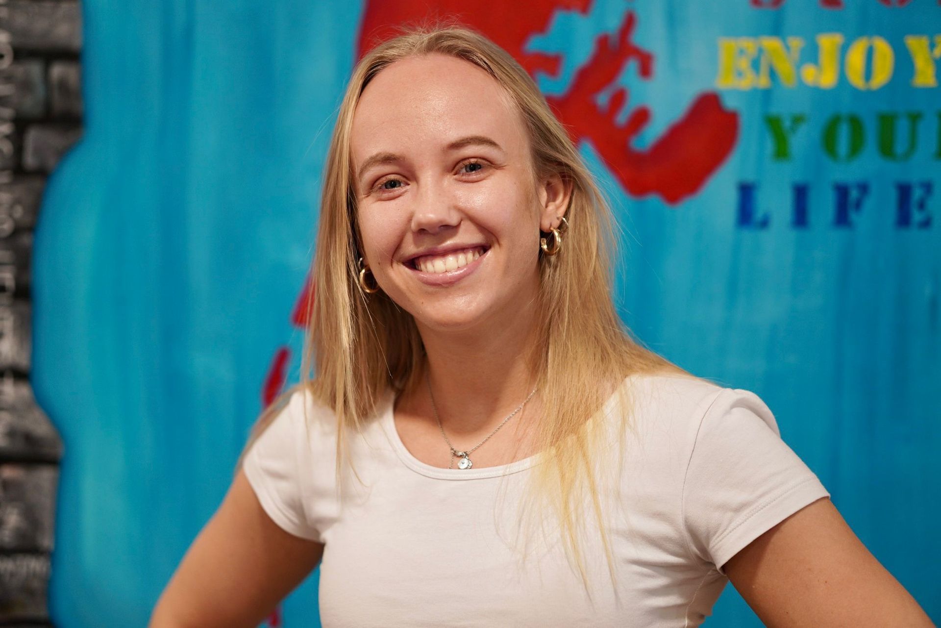 Eine Frau im weißen Hemd lächelt vor einem Schild mit der Aufschrift „Genieße dein Leben“.