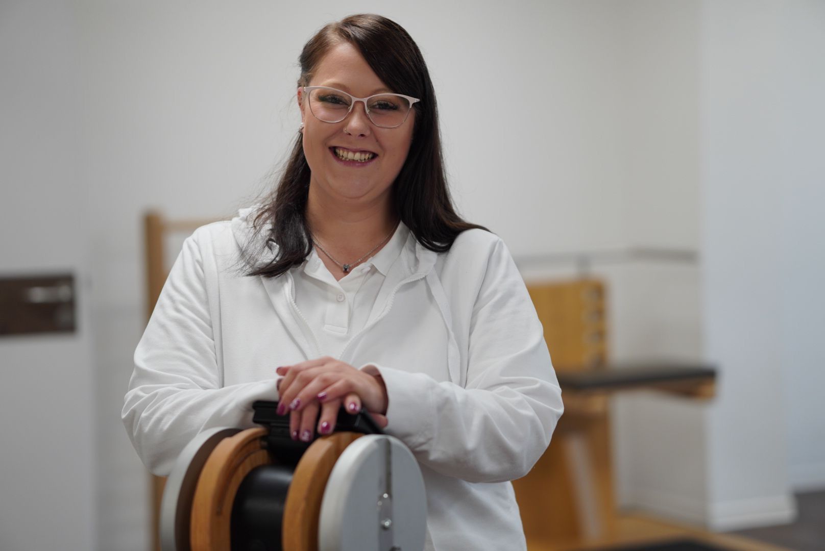 Eine Frau im weißen Laborkittel steht neben einer Maschine und lächelt.