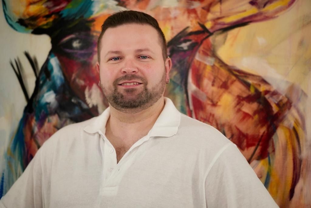 Ein Mann mit Bart steht vor einem bunten Gemälde.