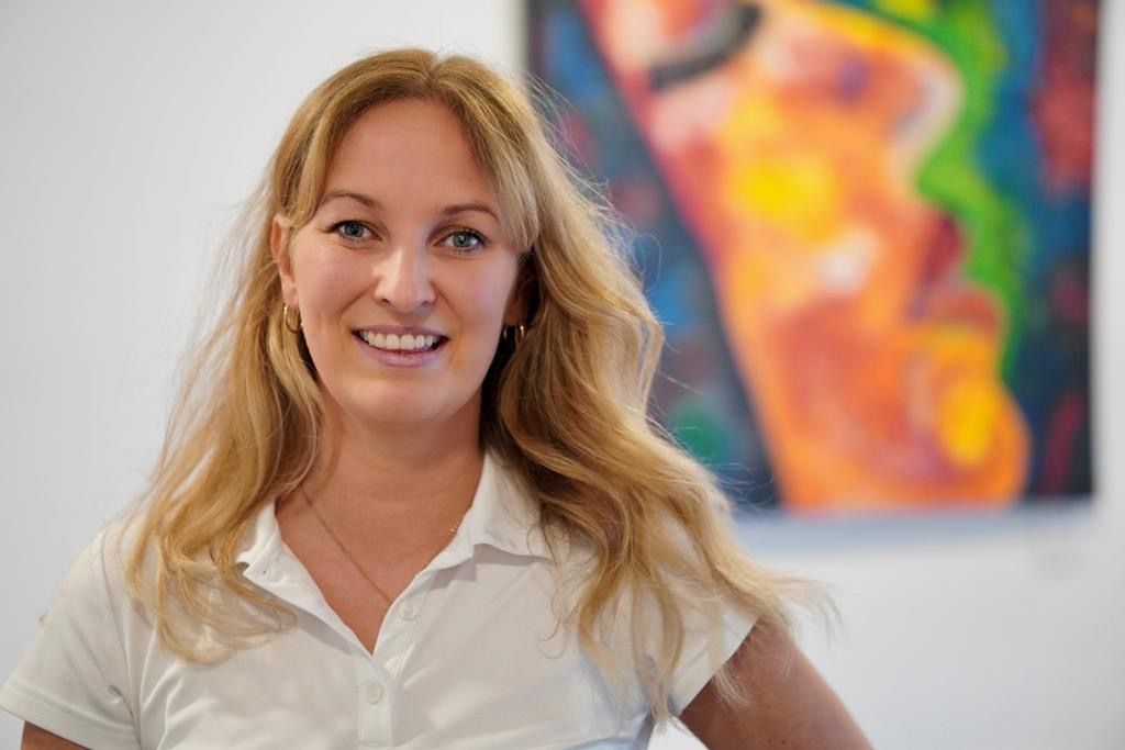 Eine Frau lächelt vor einem Gemälde mit einem Frauengesicht.