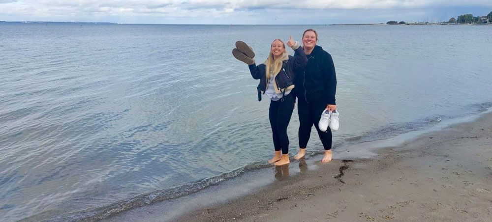 Zwei Menschen am Strand, einer winkt mit Stiefeln, der andere hält Schuhe. Grauer Himmel, Meereshintergrund.