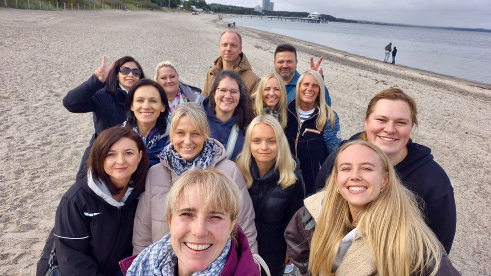 Gruppe lächelnder Menschen an einem Strand; bewölkter Himmel, Ozean im Hintergrund.