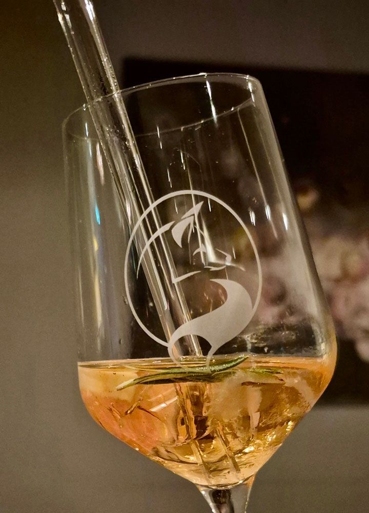 Weinglas mit rosa Getränk, Eis, Rosmarinzweig und Glasstrohhalm; auf dem Glas befindet sich ein Logo.