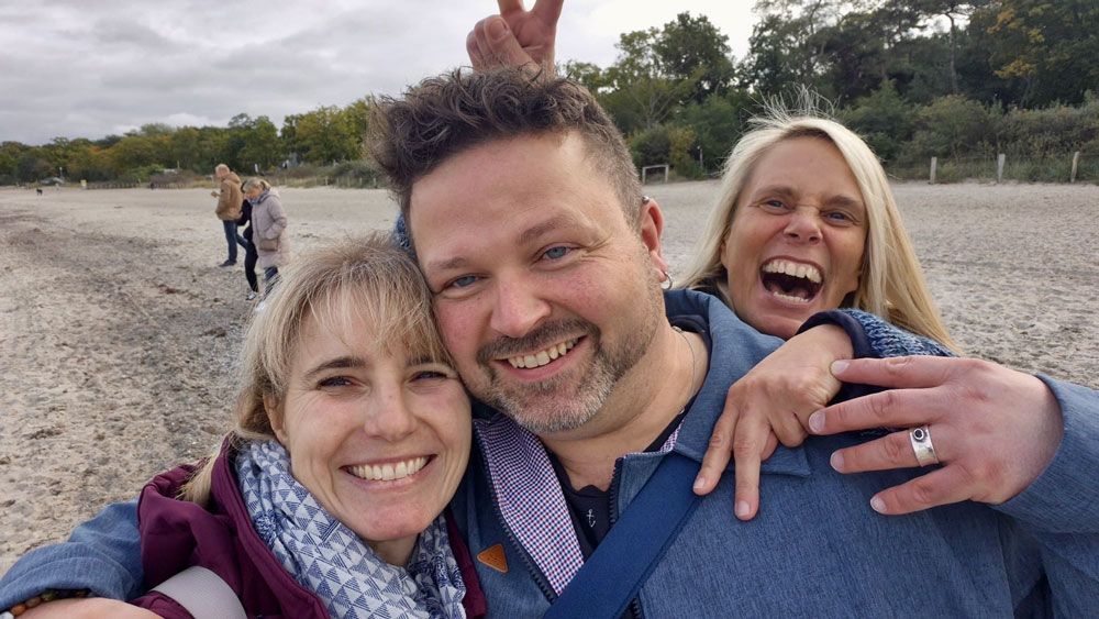 Drei lächelnde Menschen an einem Strand; in der Mitte ein Mann, auf beiden Seiten eine Frau, eine macht ein Peace-Zeichen.