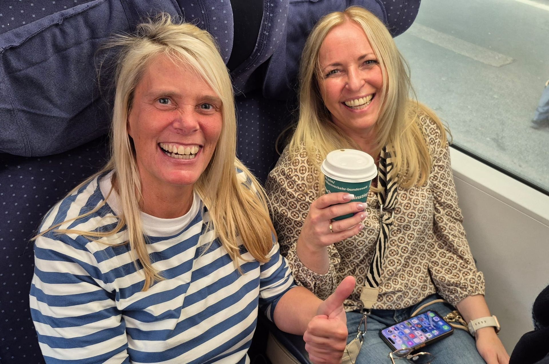 Zwei lächelnde Frauen in einem Zug; eine hält einen Kaffee und zeigt den Daumen nach oben.