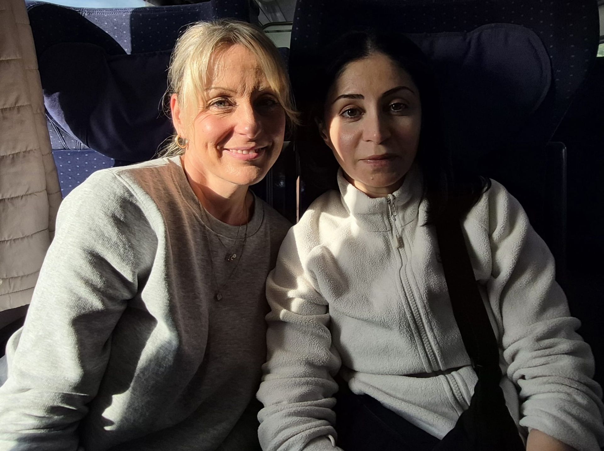 Zwei Frauen sitzen eng beieinander im Innenbereich; eine mit blonden Haaren lächelt, die andere mit Brünette ist neutral;