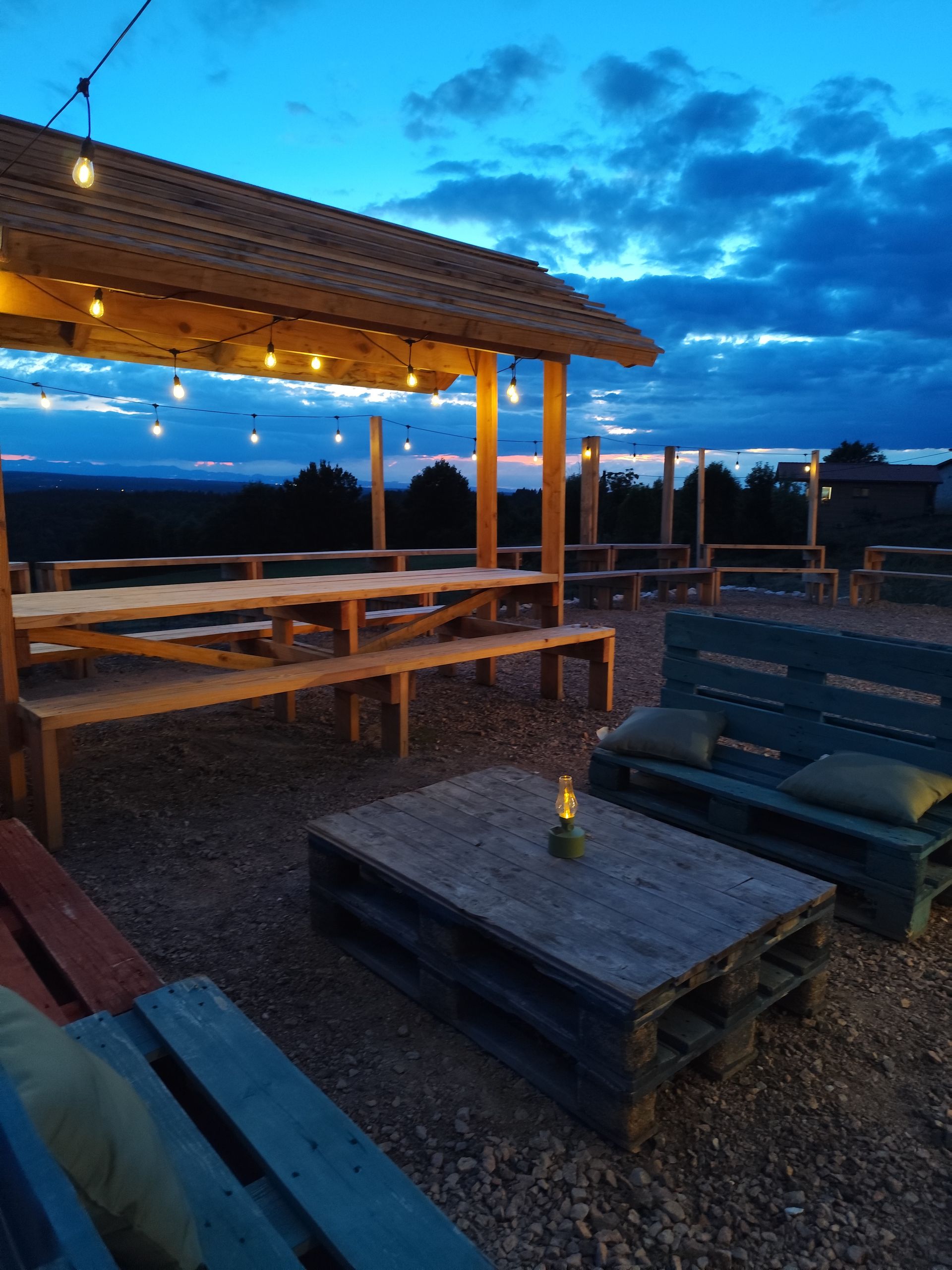 Terrasse extérieure avec guirlandes lumineuses, tables en bois et bancs au crépuscule, donnant sur un ciel d'un bleu profond.
