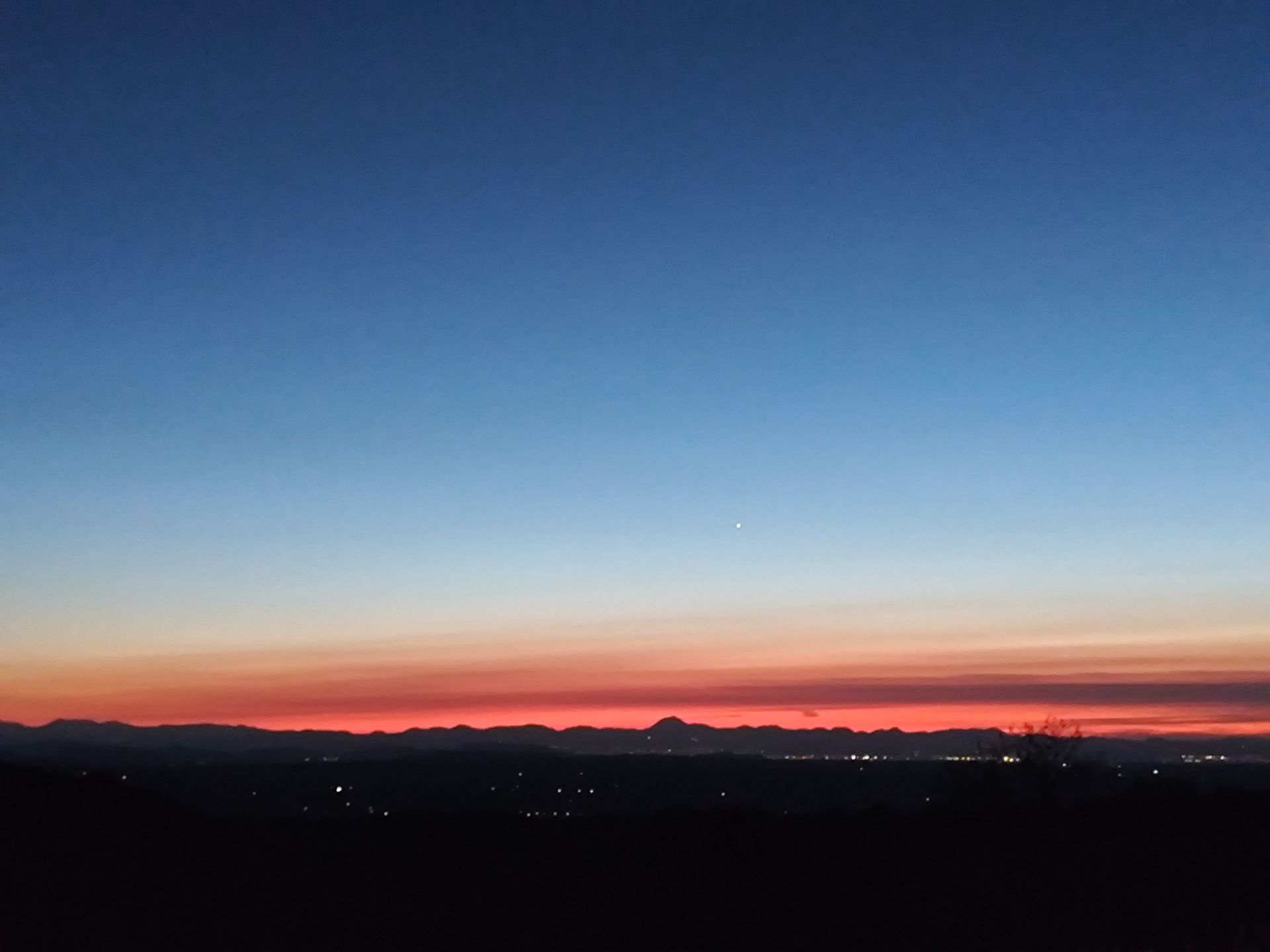 Coucher de soleil sur des montagnes lointaines avec un premier plan sombre et un ciel bleu orangé.